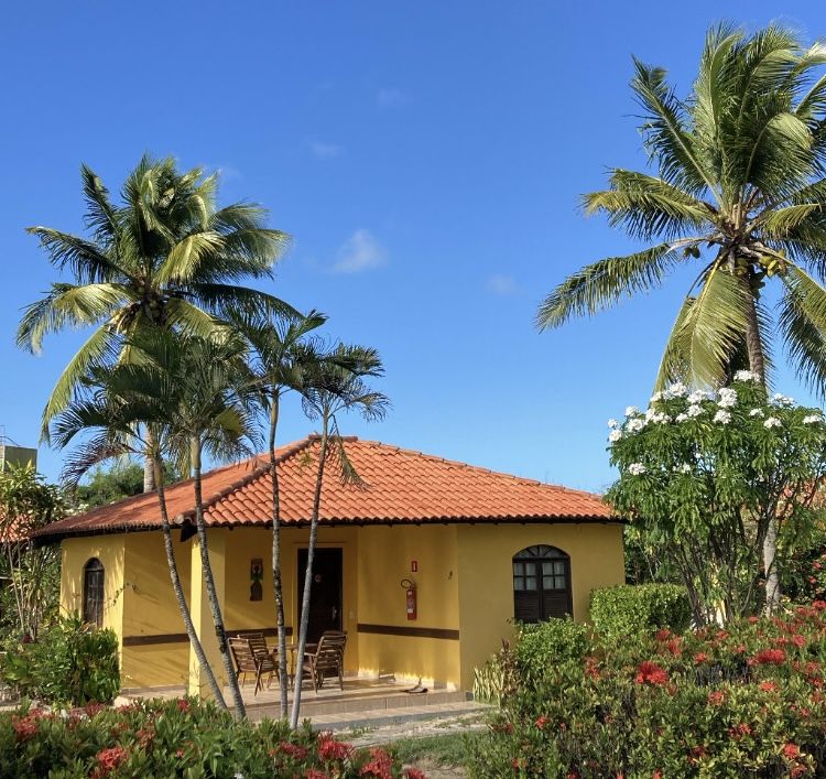 Bangalô amarelo com telhado de cerâmica entre coqueiros e jardim florido, com cadeiras na varanda
