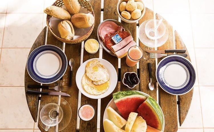 Café da manhã servido na mesa de madeira: pães, frios, ovos, frutas e suco