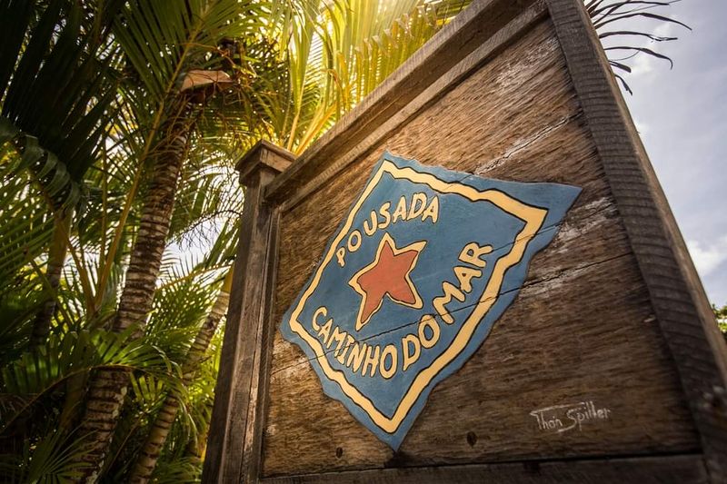 Placa de madeira na entrada com a logo pintada à mão: losango azul, estrela vermelha e o nome da pousada