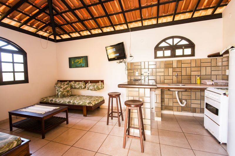 Sala e cozinha integradas do bangalô, com sofá-cama, mesa de centro de madeira e telhado aparente