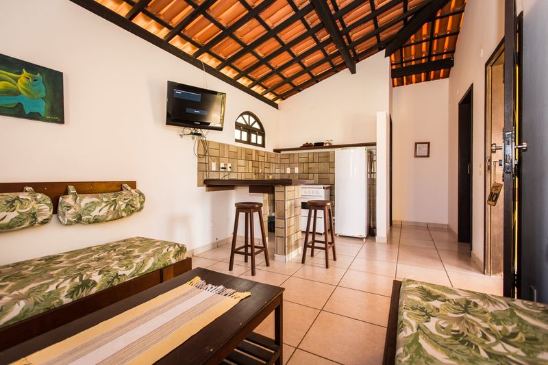 Sala do bangalô com sofás-cama e TV, integrada à cozinha com bancada, banquetas, fogão e geladeira
